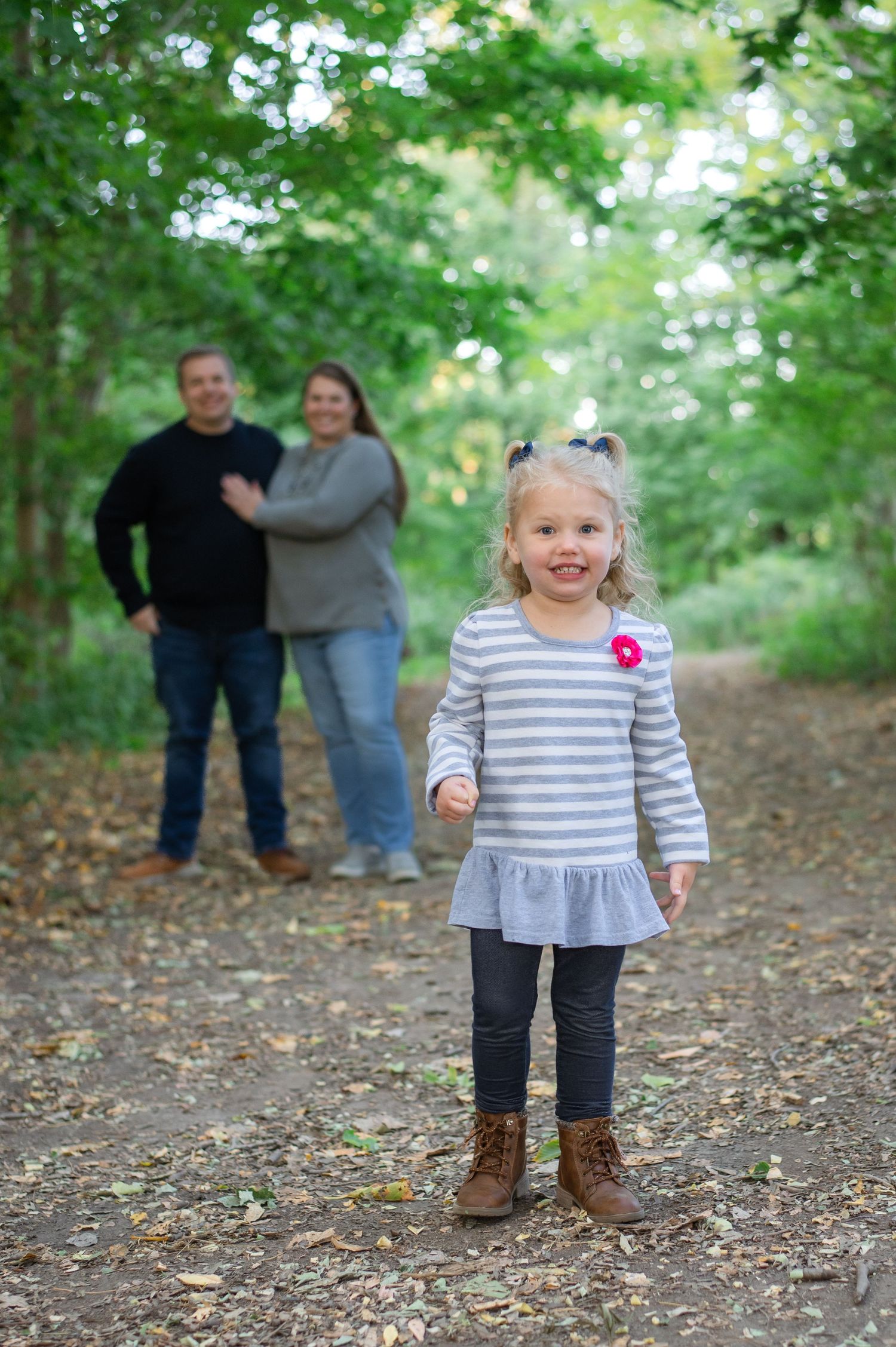 A series of photos showing a toddler in striped clothing and boots playing on a leaf-covered forest path while adults watch.