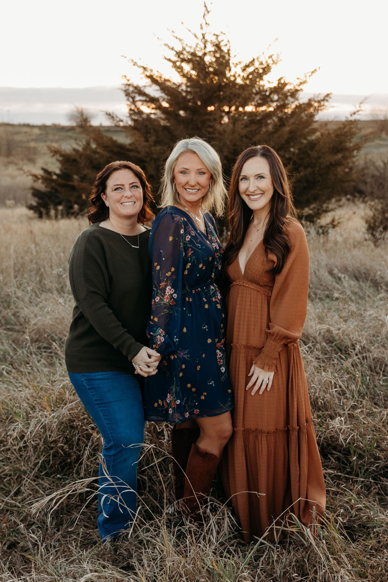Three friends in casual fall outfits laugh together in a field at sunset with evergreen trees in the background.