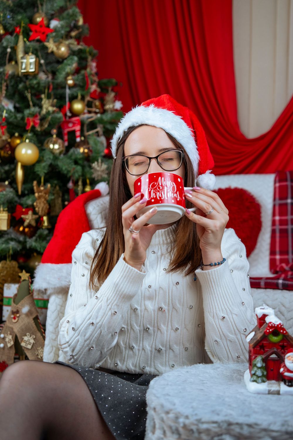 Someone wearing a Santa hat and white sweater drinks from a festive red mug by a decorated Christmas tree and red curtain.