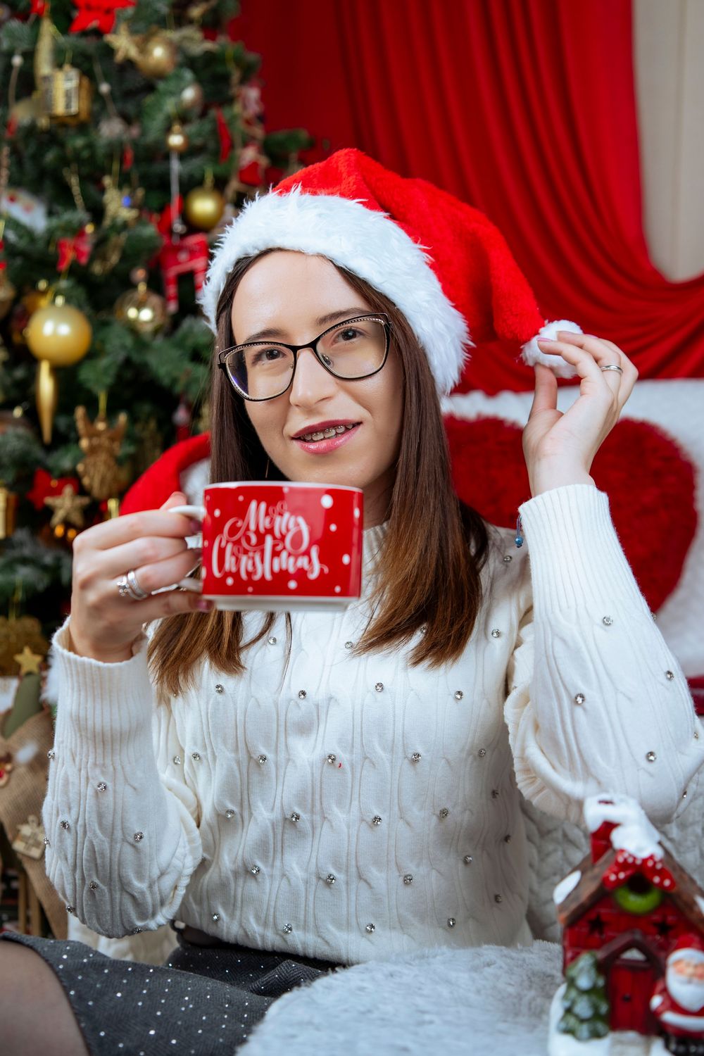 Person in white knit sweater and Santa hat holds festive red coffee mug against Christmas tree background.
