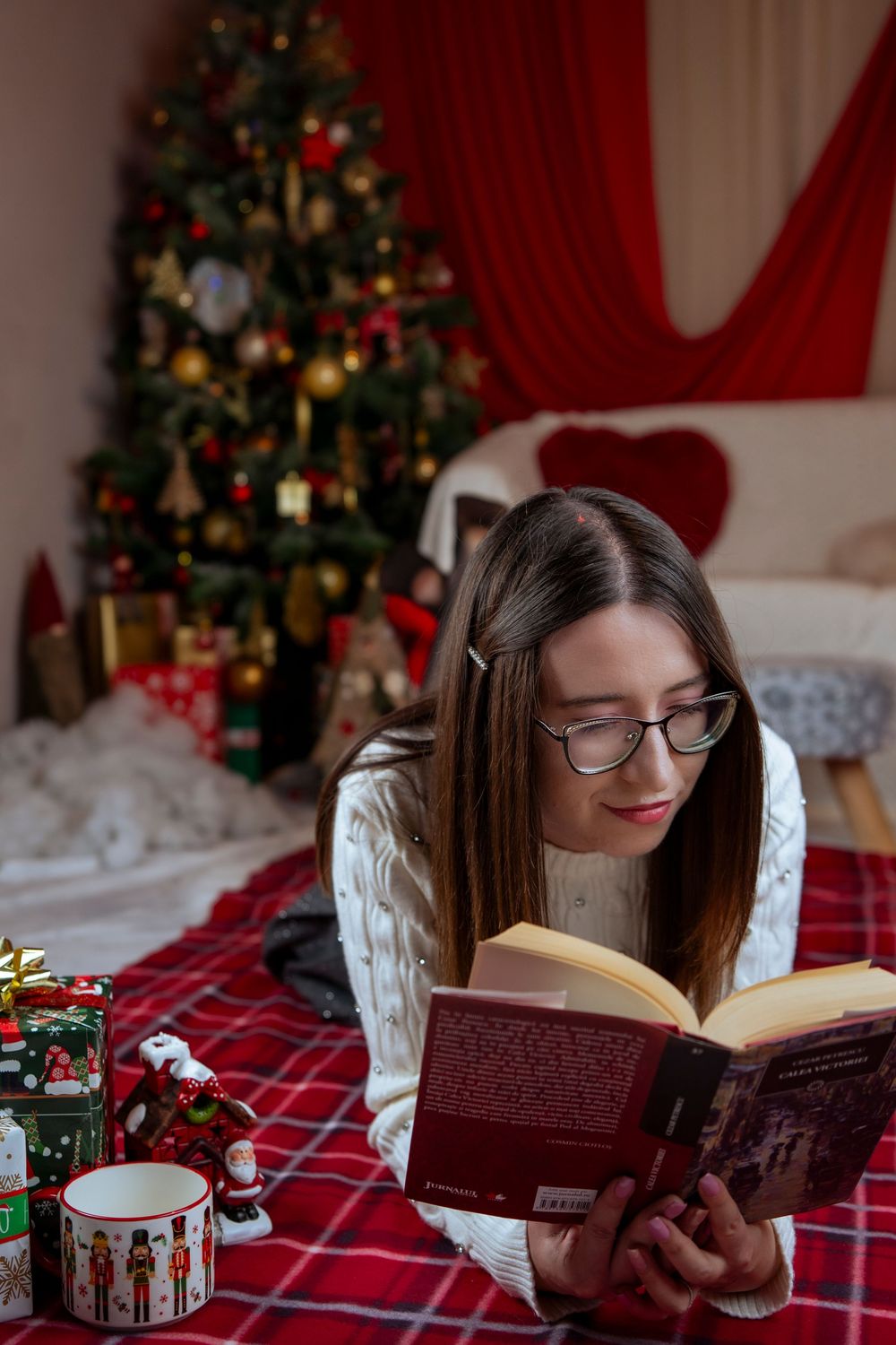 Person in cozy white sweater reads book by decorated Christmas tree with holiday mug nearby.