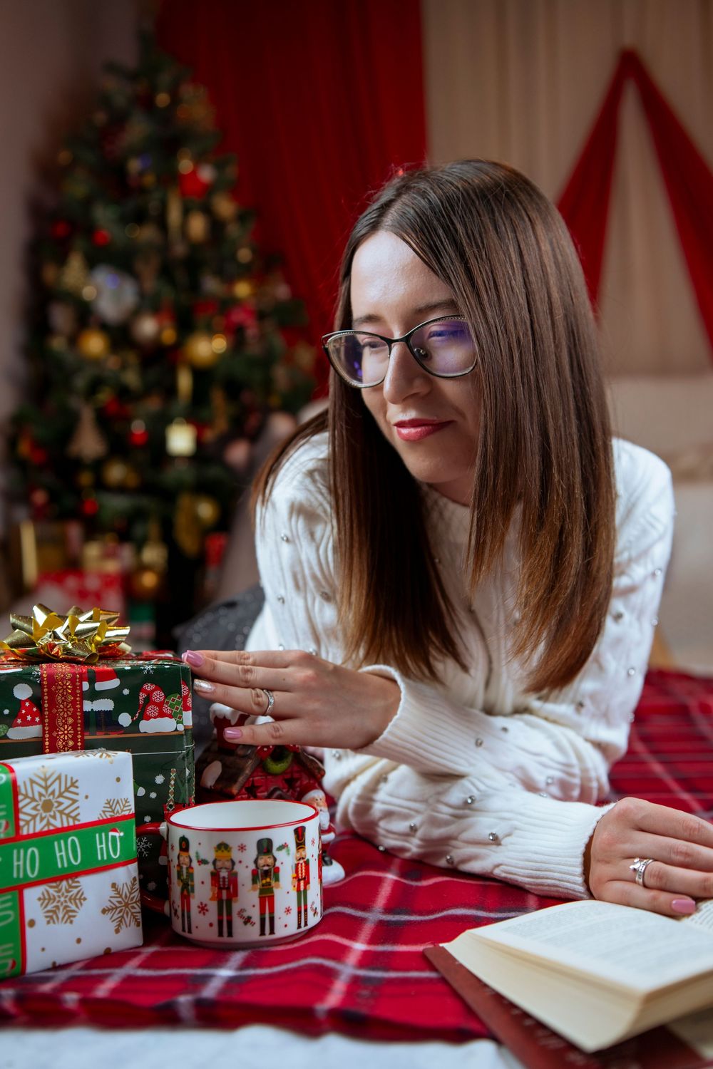 A cozy Christmas scene shows someone in a white sweater reading and wrapping gifts on a red tablecloth near a decorated tree.