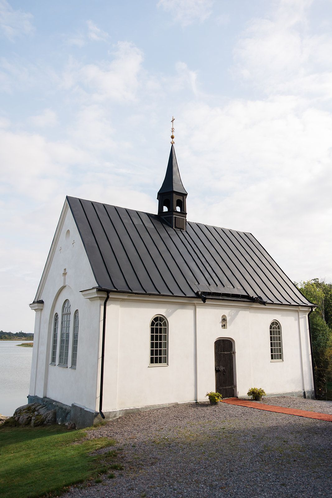 A small white church with a metal roof and arched windows sits on a gravel path against a cloudy sky.
