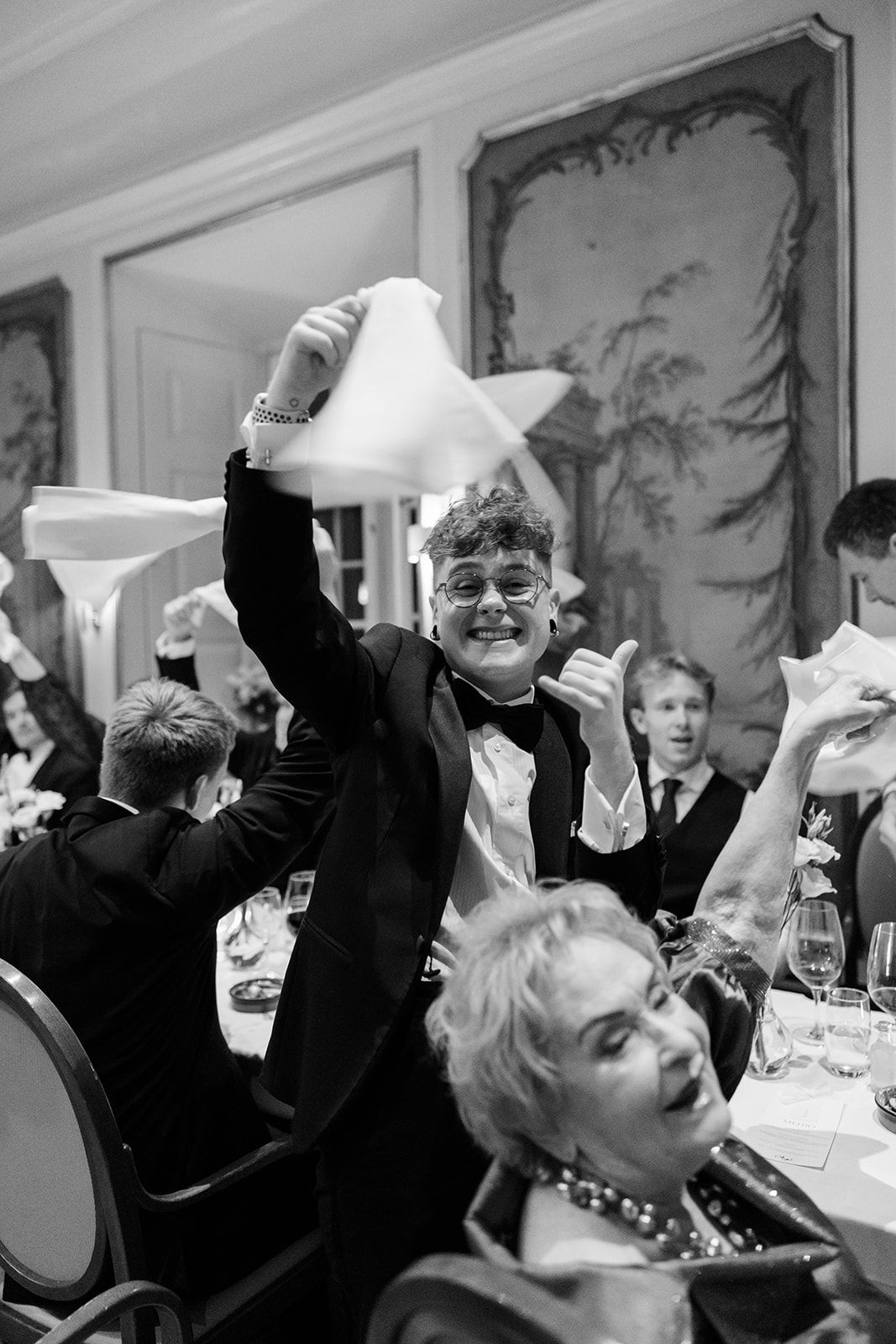 Black and white photo of guests celebrating at an elegant formal dinner party with napkin waving and lively expressions.