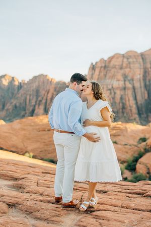 Couple in white and light blue attire embrace on red rock formations at sunset.