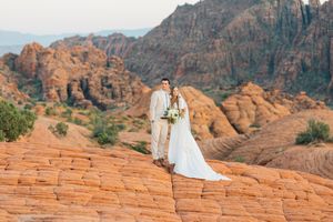 Wedding couple stands on red sandstone cliffs with dramatic mountain backdrop at sunset.