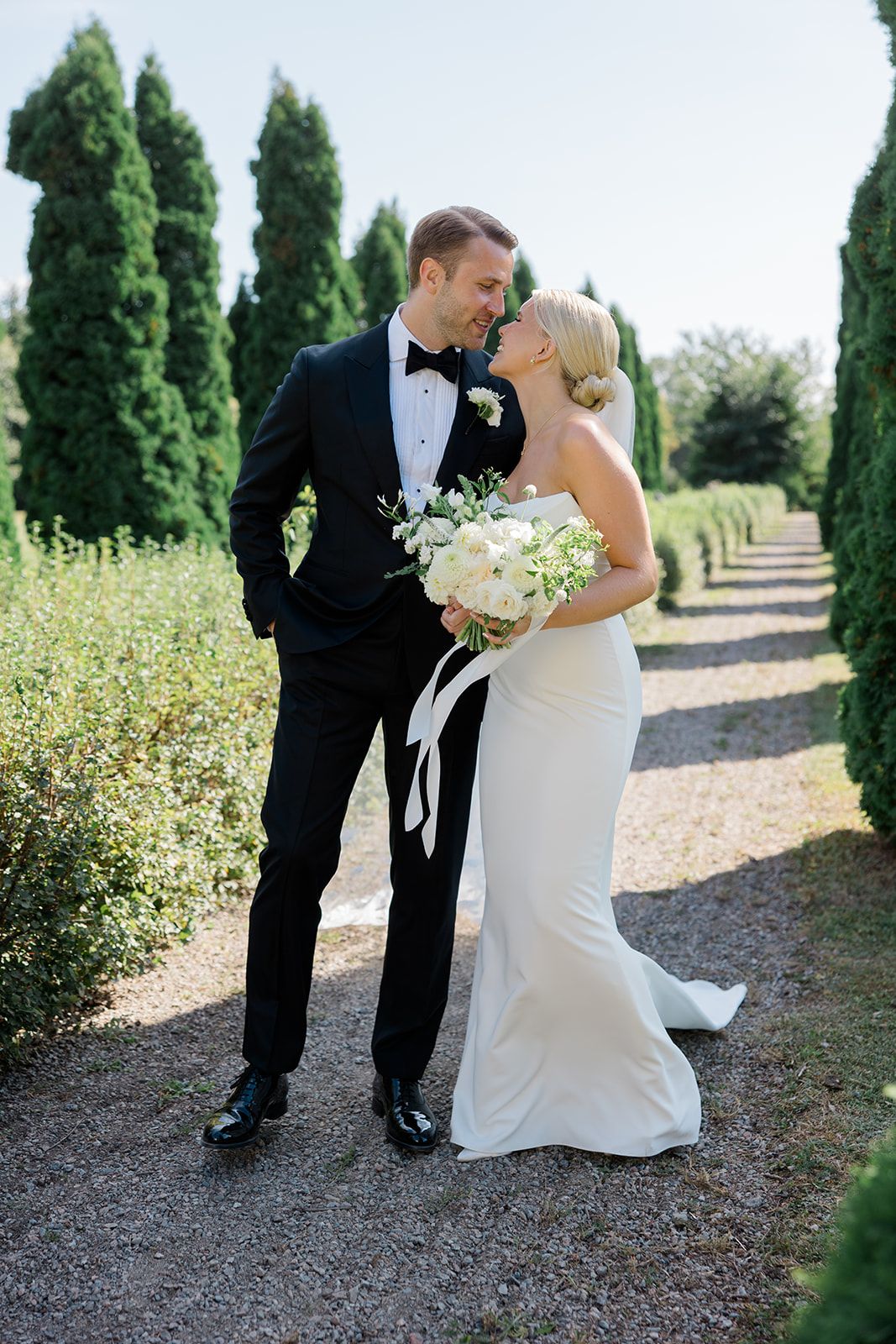 A romantic wedding couple shares an intimate moment among cypress trees lining a garden path.