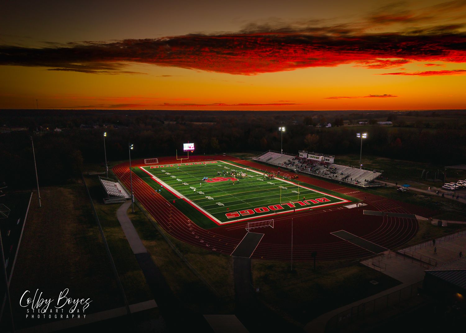 A stunning panoramic view of a high school football stadium at sunset with vibrant orange and red skies.