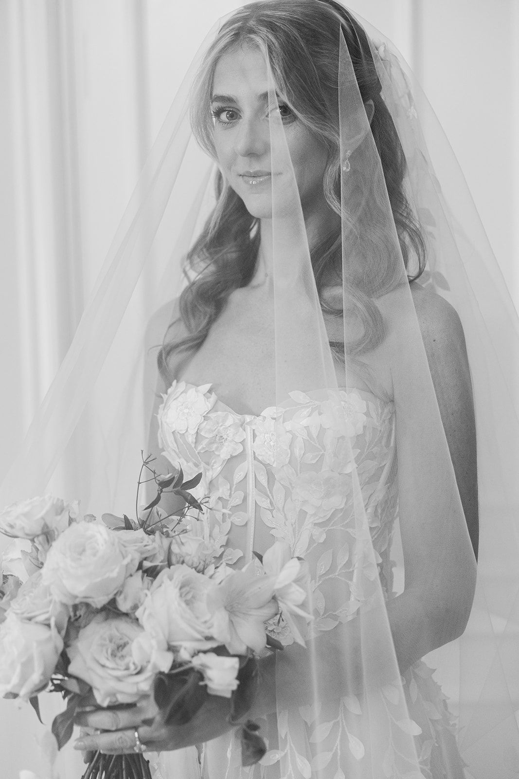 Black and white bridal portrait showing a bride in a lace dress with veil holding a rose bouquet.