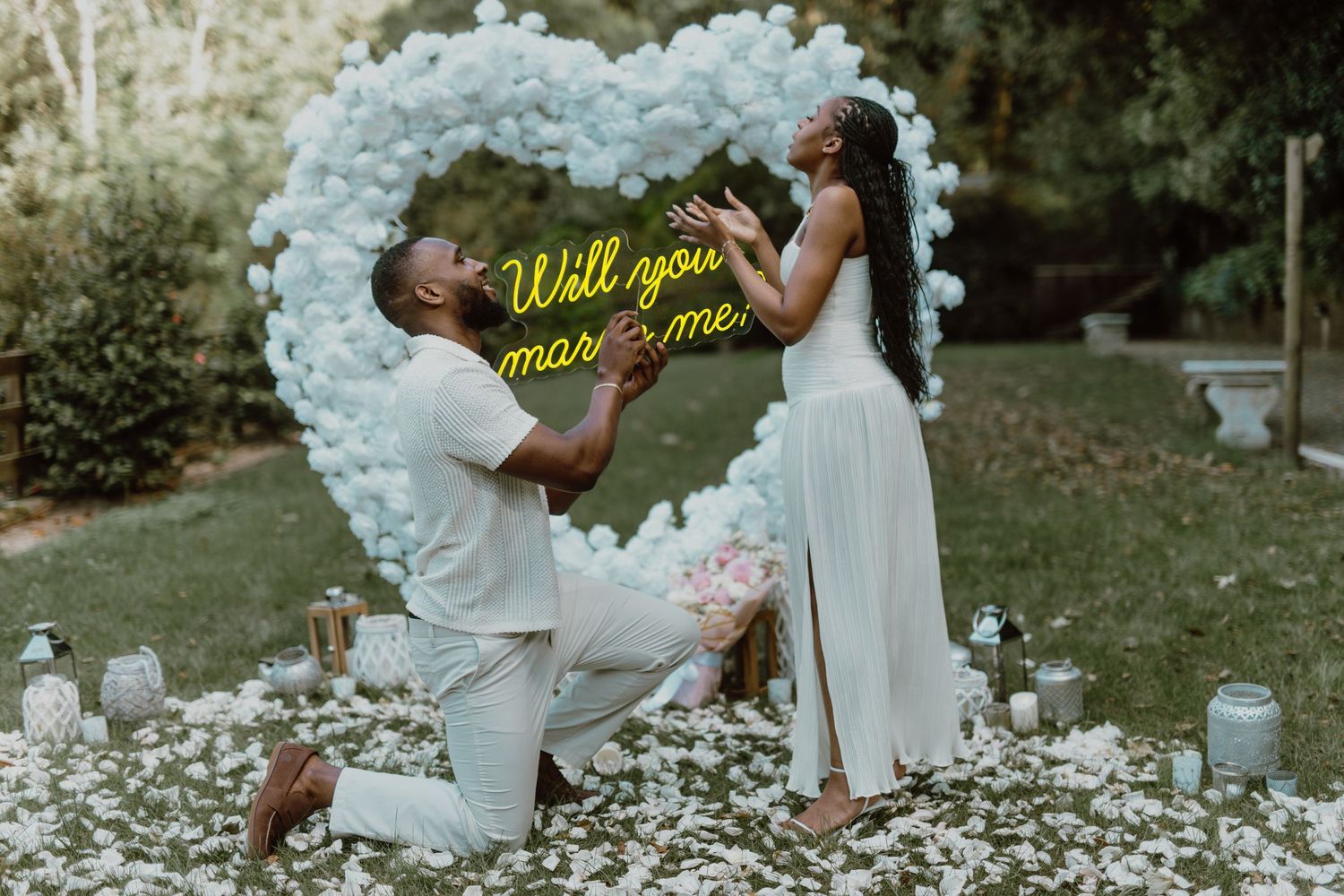 Romantic marriage proposal scene with illuminated heart-shaped balloon arch and rose petals scattered on grass.