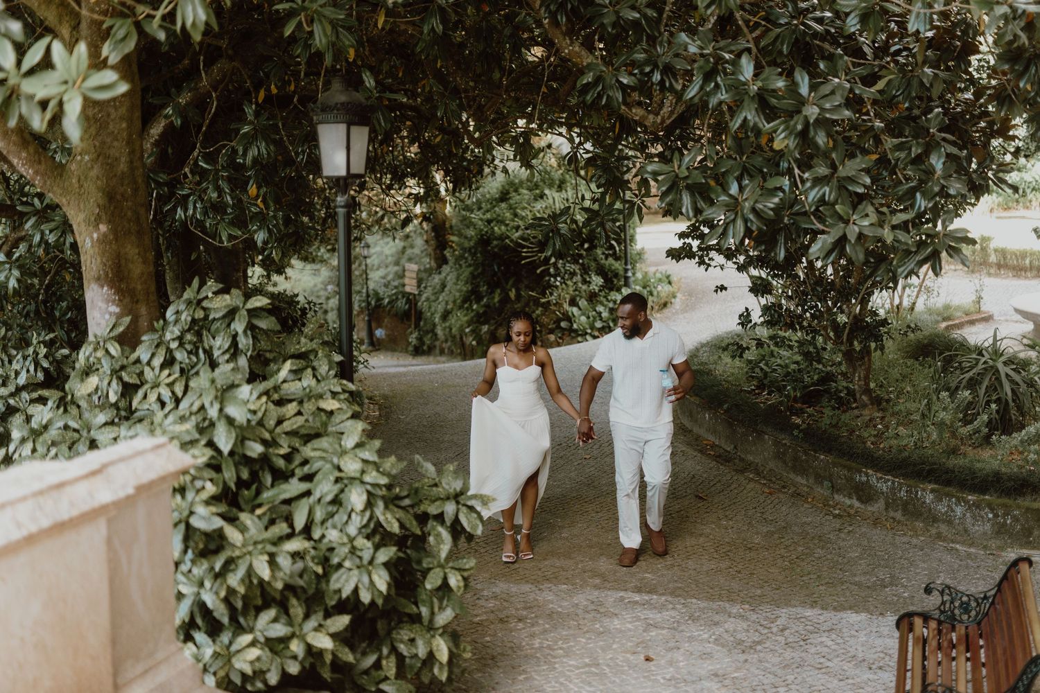 A couple walks hand in hand down a garden path surrounded by lush greenery and vintage lamp posts.
