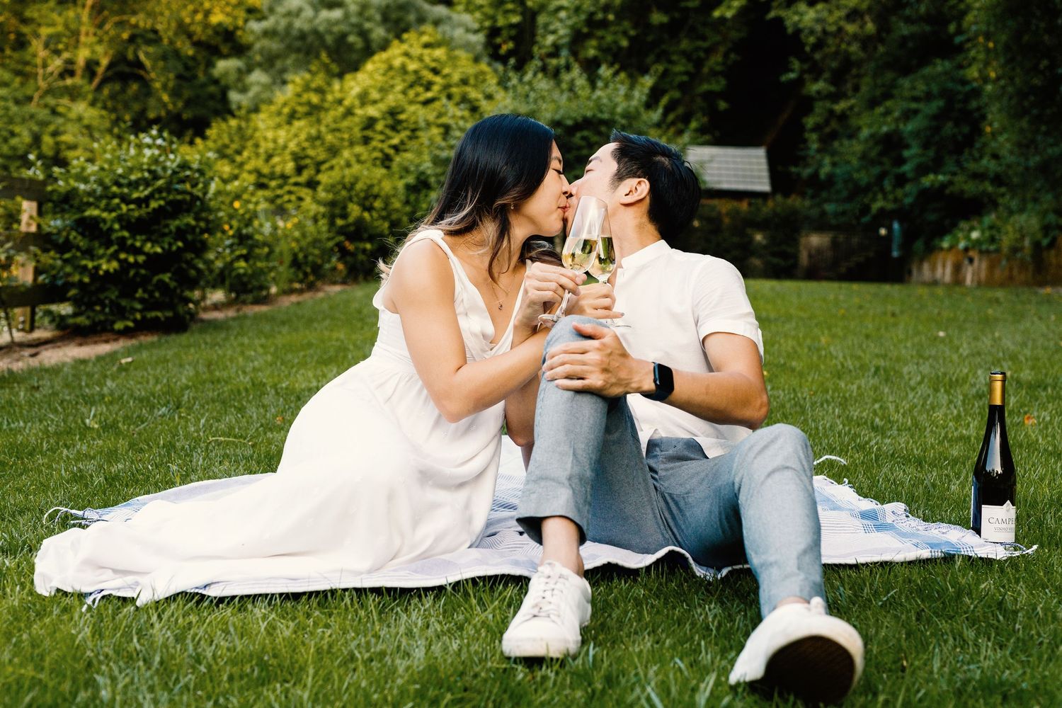 Couple enjoying wine and sharing a romantic picnic together on a grassy lawn in a garden setting.