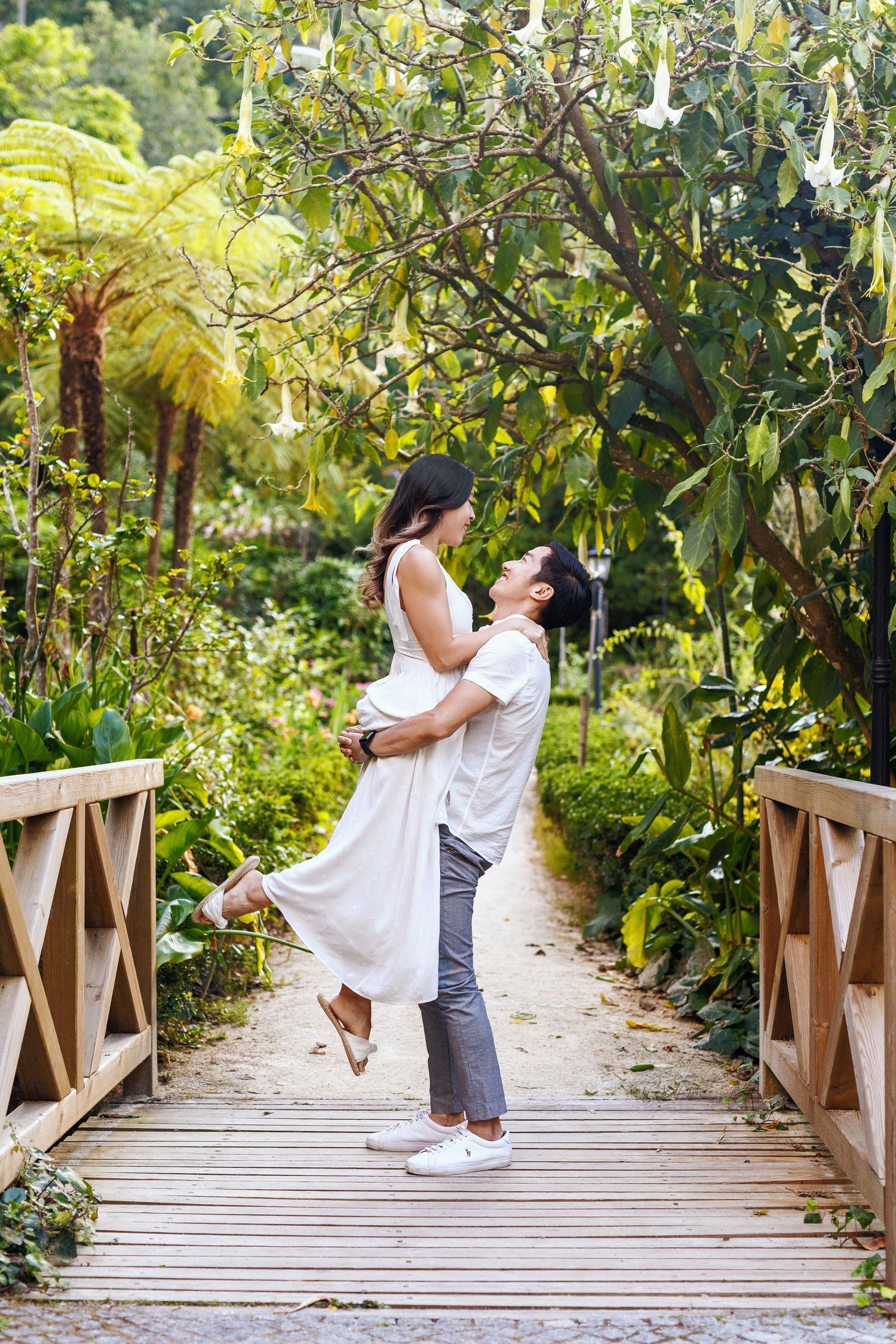A couple embraces on a wooden footbridge surrounded by lush tropical garden foliage and ferns.