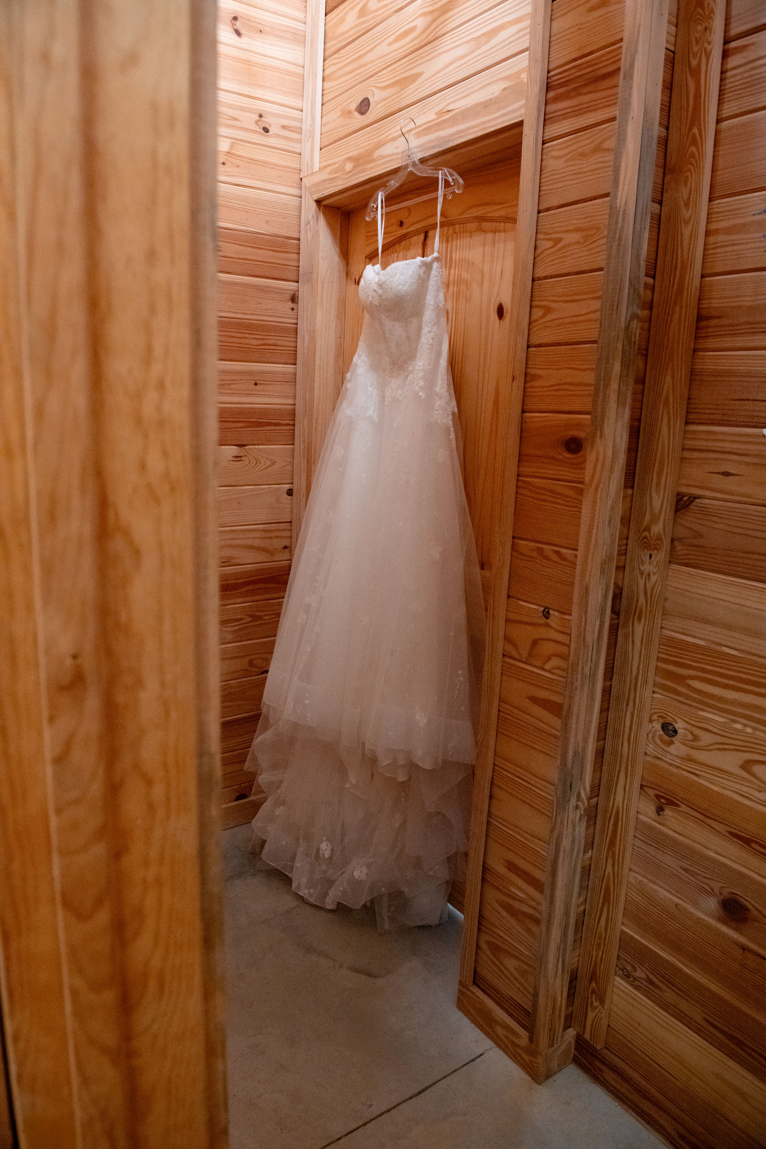 A white wedding dress hangs elegantly against rustic wooden walls in a bridal preparation room.