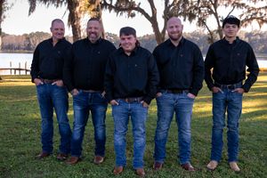 Five people wearing similar dark shirts and blue jeans stand together outdoors by a lake at sunset. Groom and groomsmen.