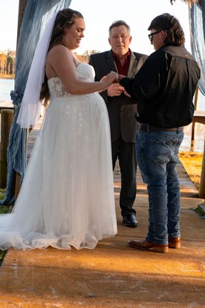 A bride in a white wedding dress stands with others during an outdoor sunset ceremony.