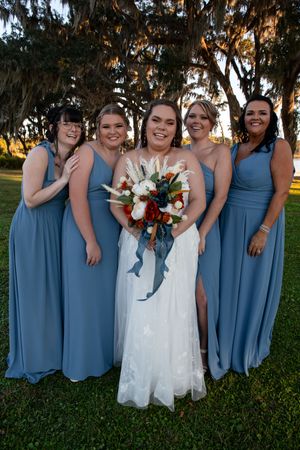 Wedding party in dusty blue bridesmaid dresses poses together on a grassy lawn at sunset.