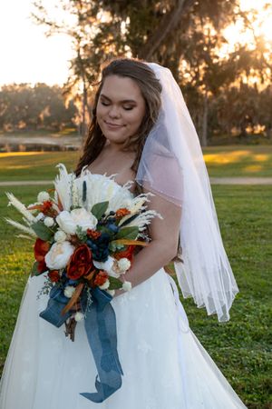 A bride in a white ballgown holds a vibrant autumn bouquet with red flowers and blue ribbons at sunset.