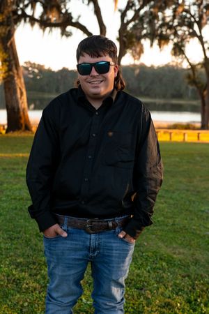 A groom in sunglasses and casual attire poses outdoors at sunset with trees in the background.