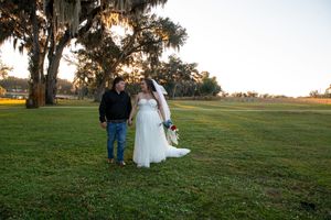 A couple stands together on a grassy field at sunset with Spanish moss hanging from trees.