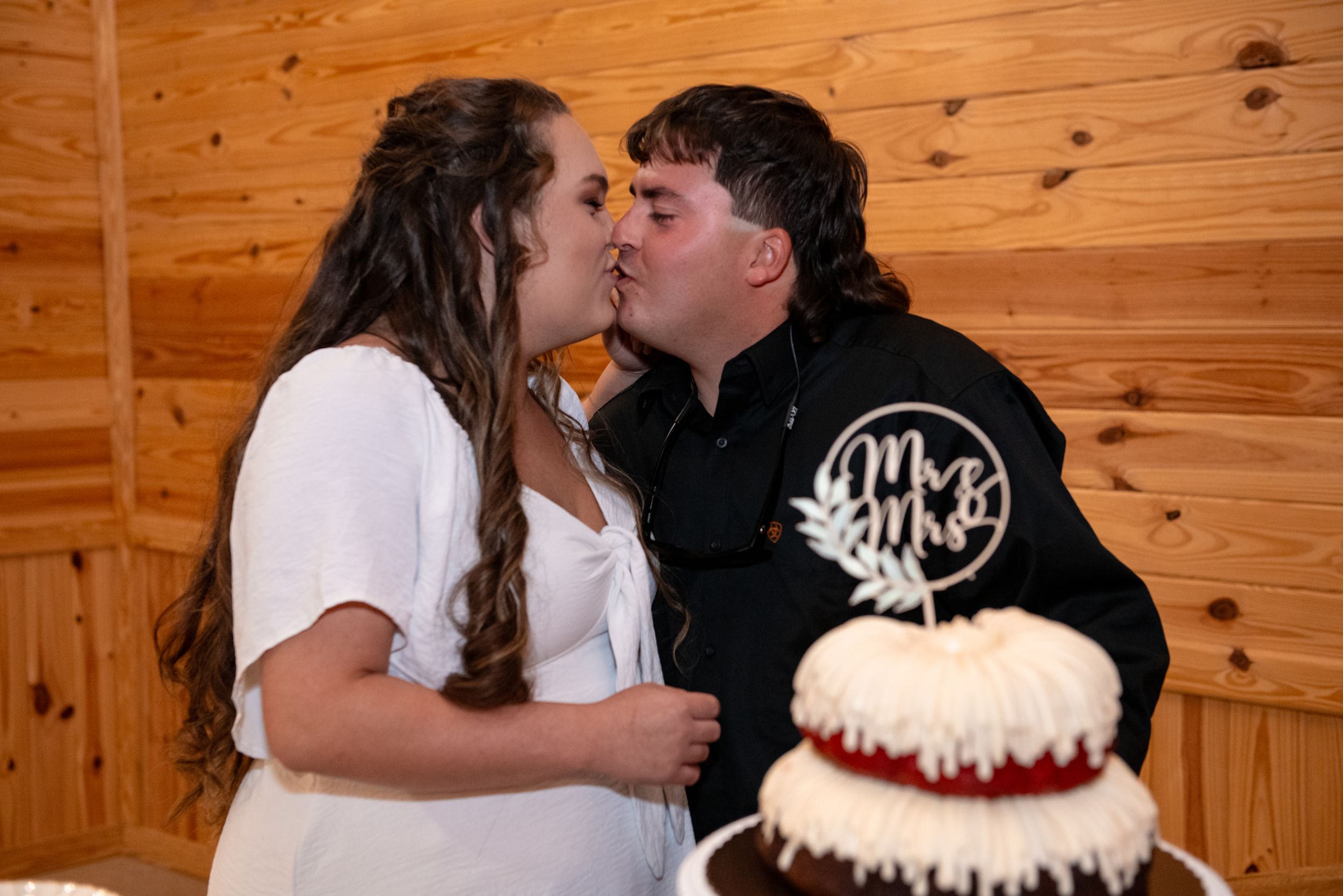 A couple shares a kiss behind their two-tiered wedding cake in a rustic wooden venue.