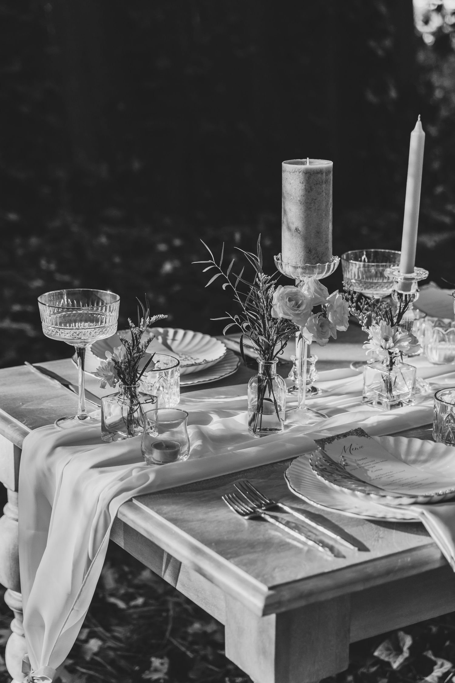 Elegant black and white table setting with white linens, crystal glasses and floral centerpieces on rustic wooden table.