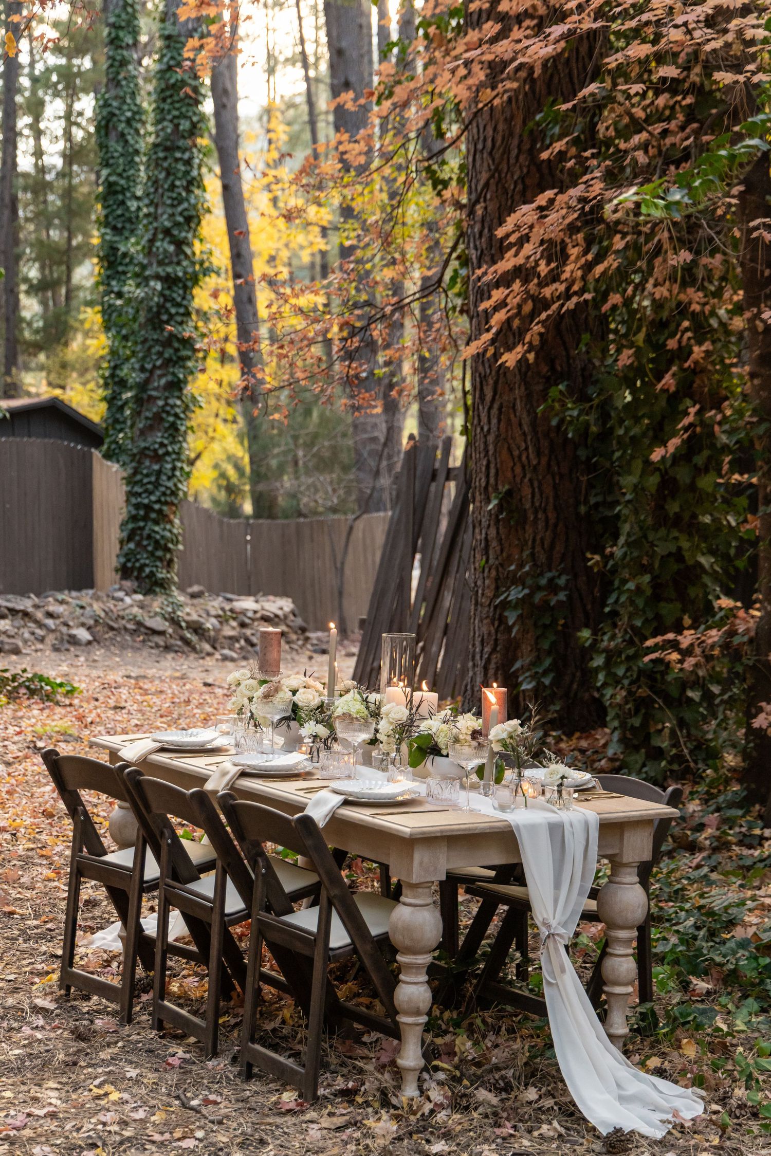 Rustic outdoor dining table with wooden chairs set up in a wooded area with autumn foliage and ivy-covered trees.