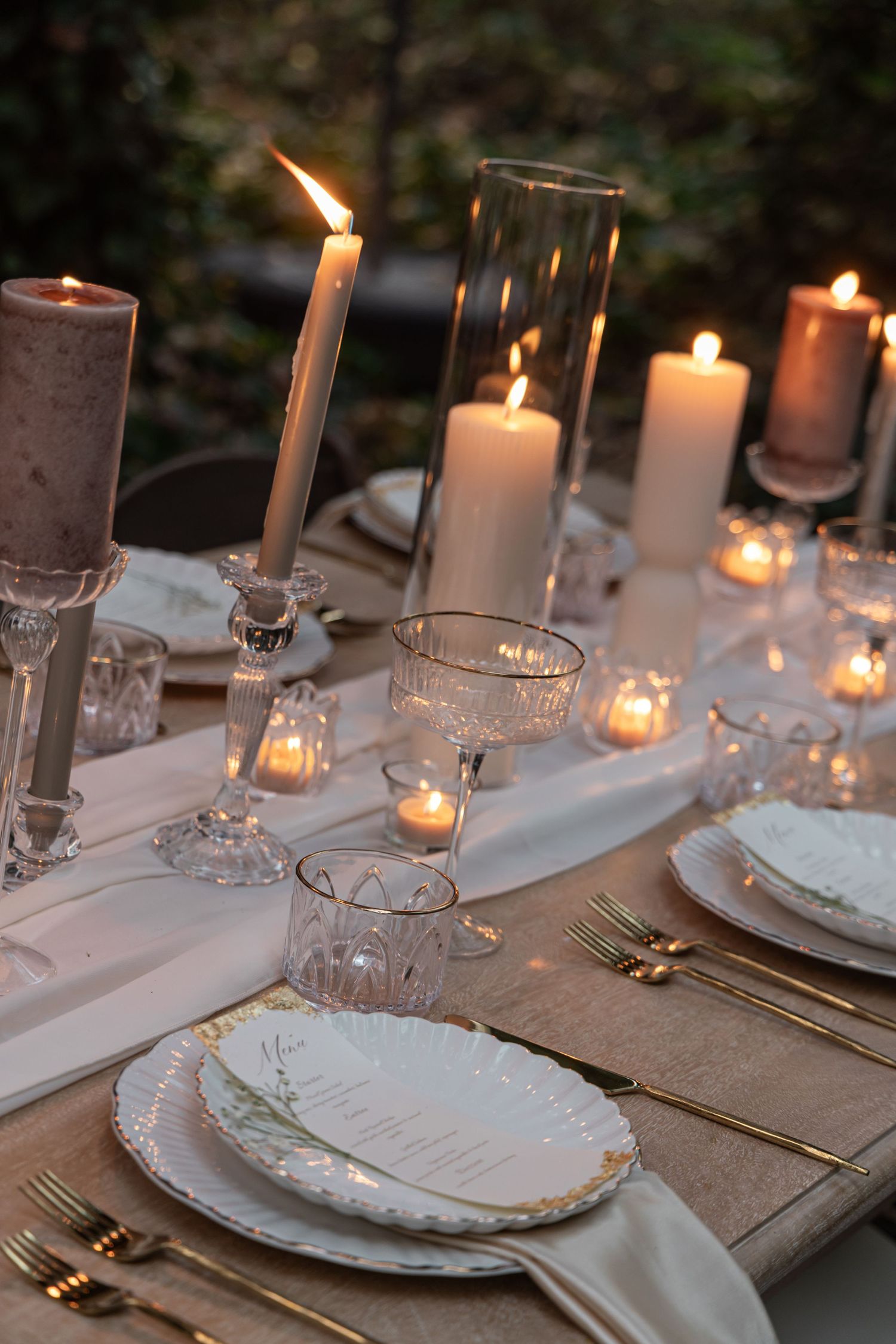 Elegant candlelit table setting with white plates and linen runner features multiple glowing tapered candles in glass holders.