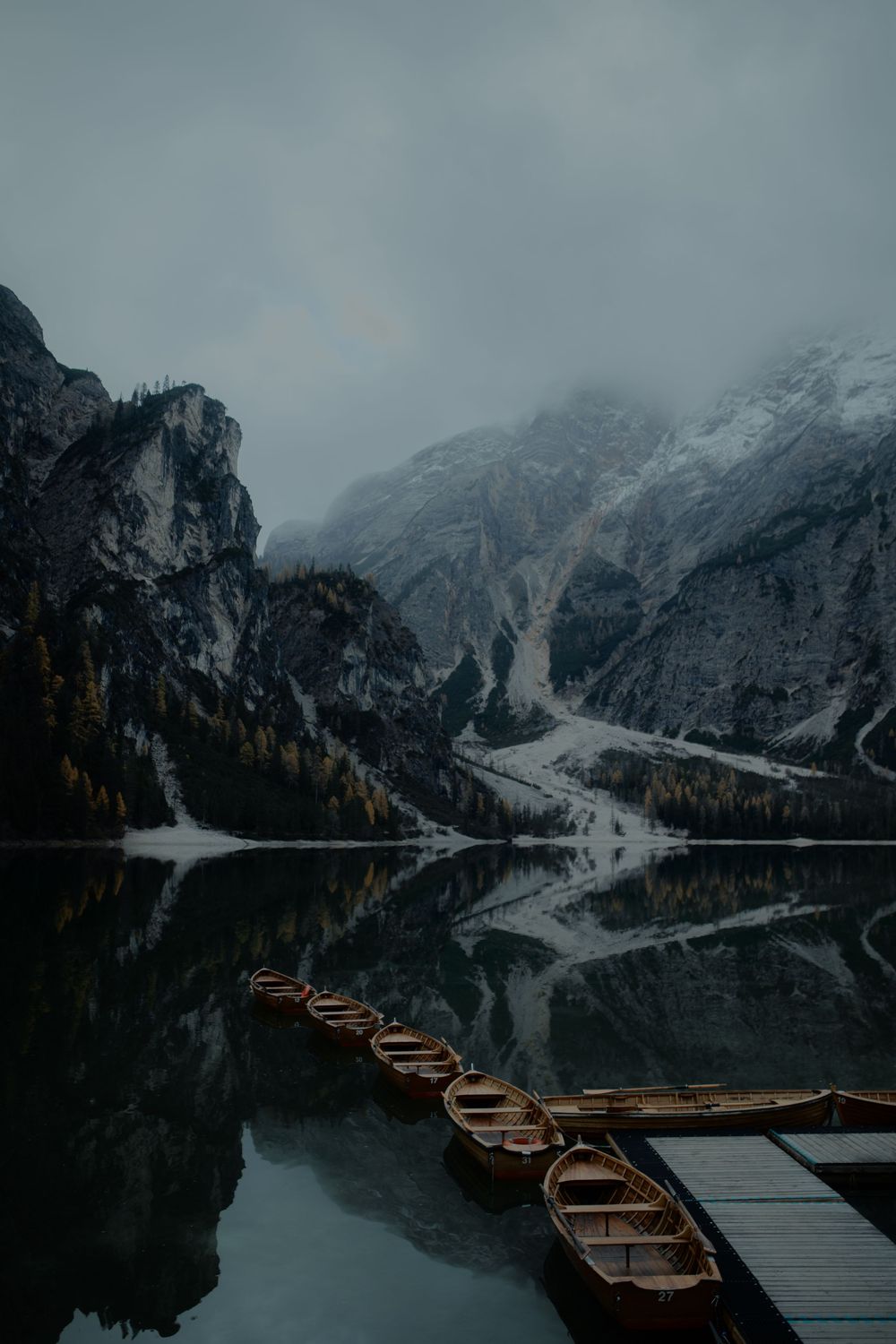 Wooden rowing boats docked at a misty mountain lake with snow-capped peaks and autumn trees reflecting in dark waters.
