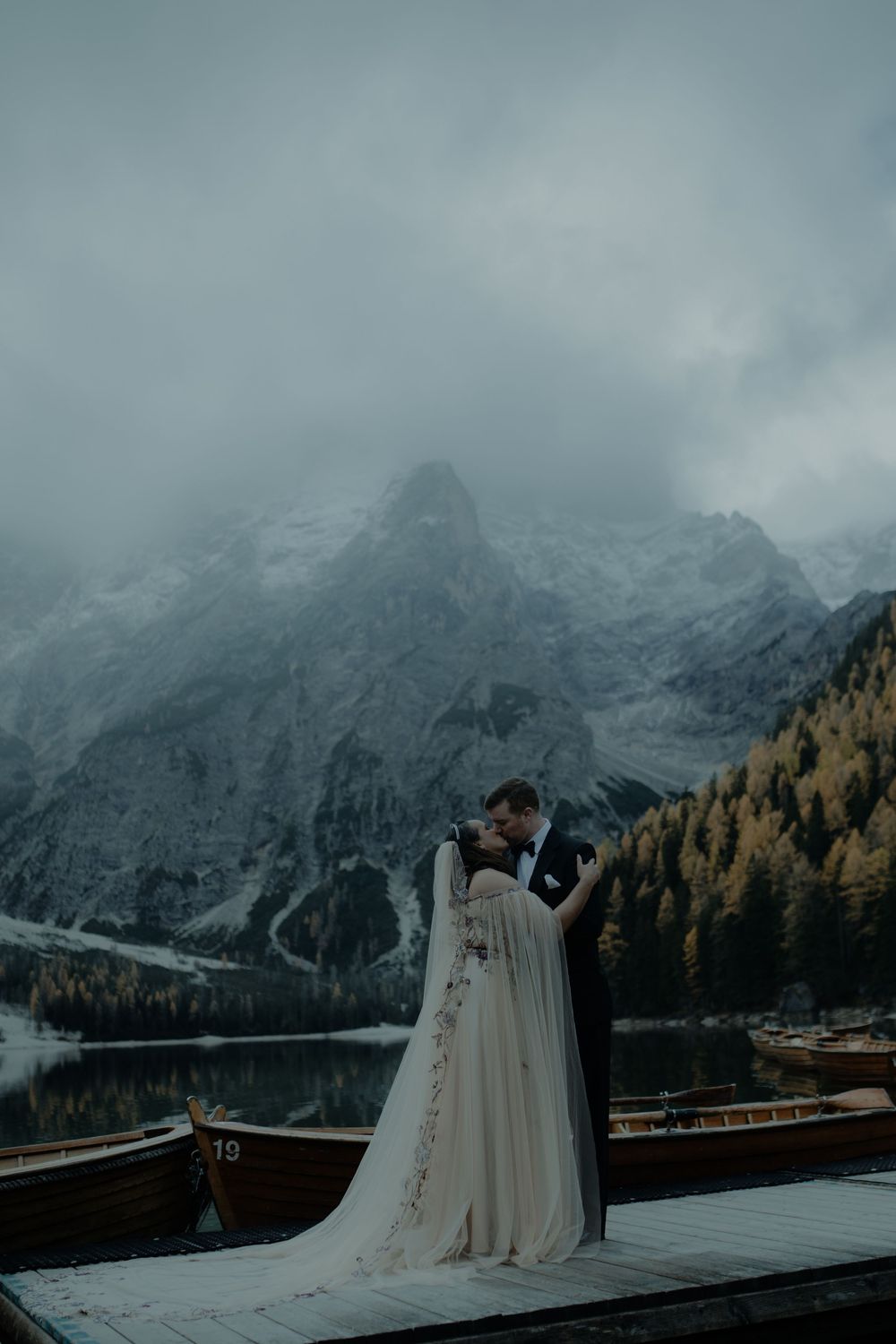 Bride and groom embrace on a dock at Lago di Braies with misty Dolomite mountains and autumn trees reflected in the lake.