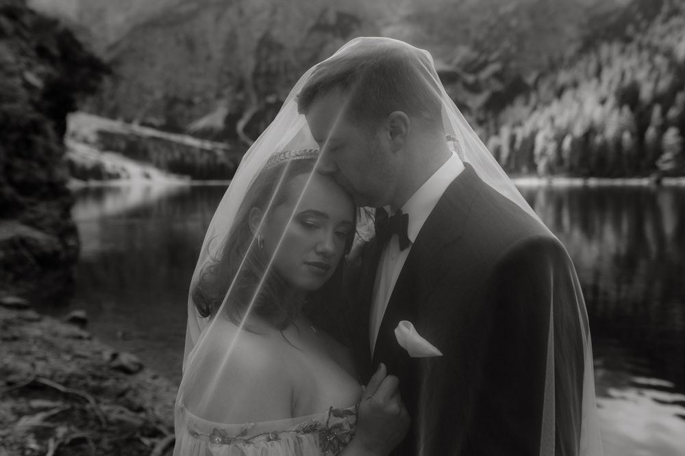 Black and white photo of bride and groom sharing intimate moment under veil by mountain lake.