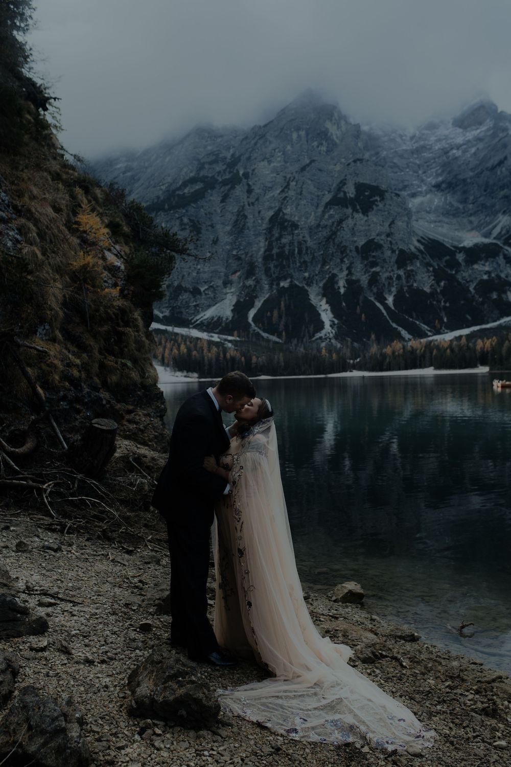 Wedding couple kissing on rocky shore of Lago di Braies with dramatic snow-dusted Dolomite mountains and calm lake reflections.