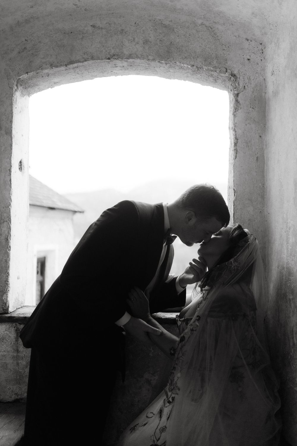Black and white photo of a bride and groom sharing a romantic kiss in an arched stone window.