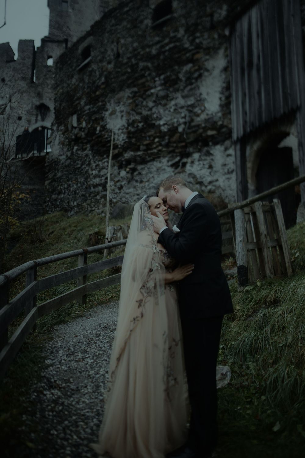A couple shares a romantic moment in their wedding attire along a rustic wooden fence at dusk.