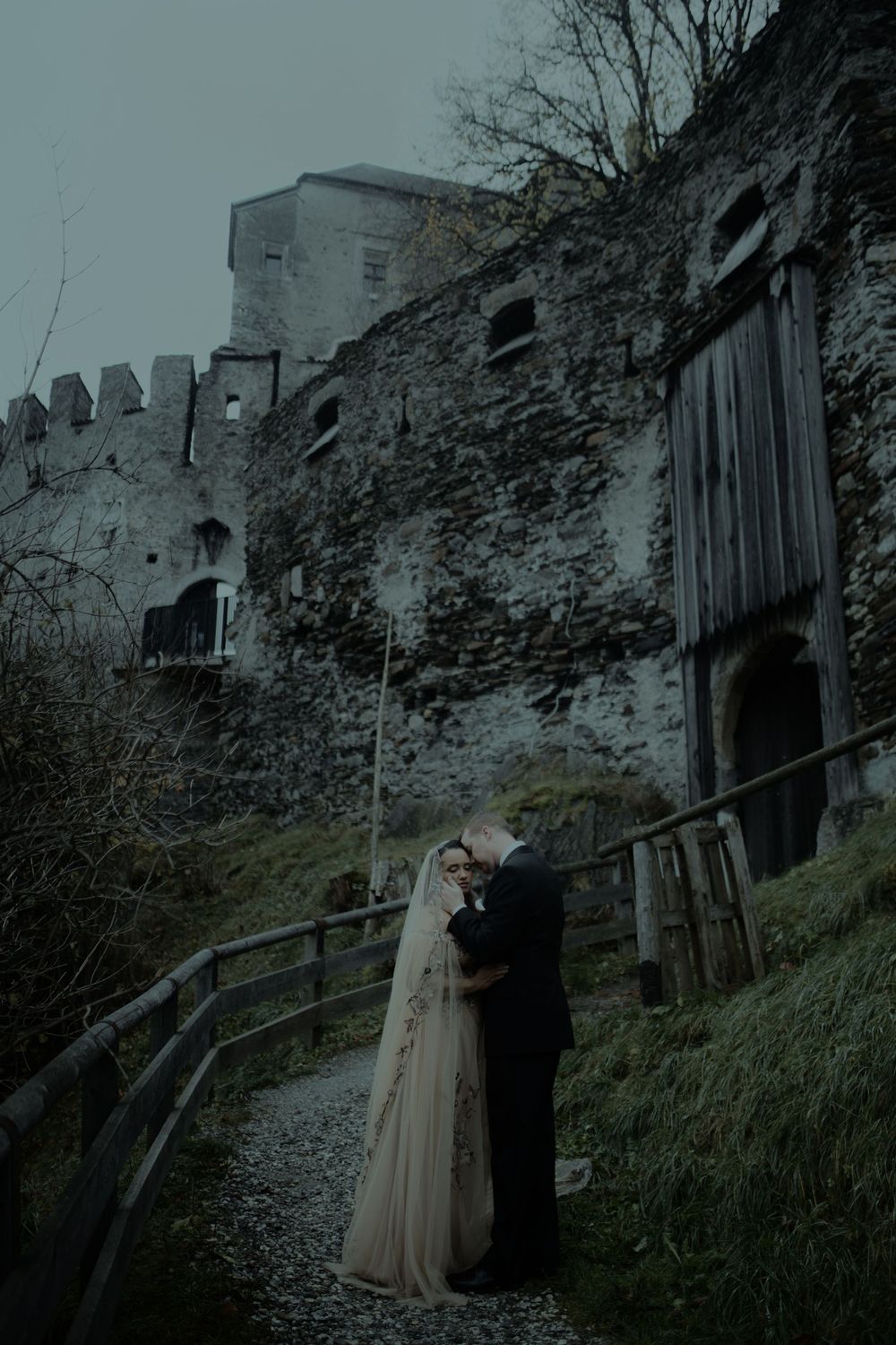 A moody wedding photoshoot against stone ruins and wooden structures in a misty, atmospheric setting.