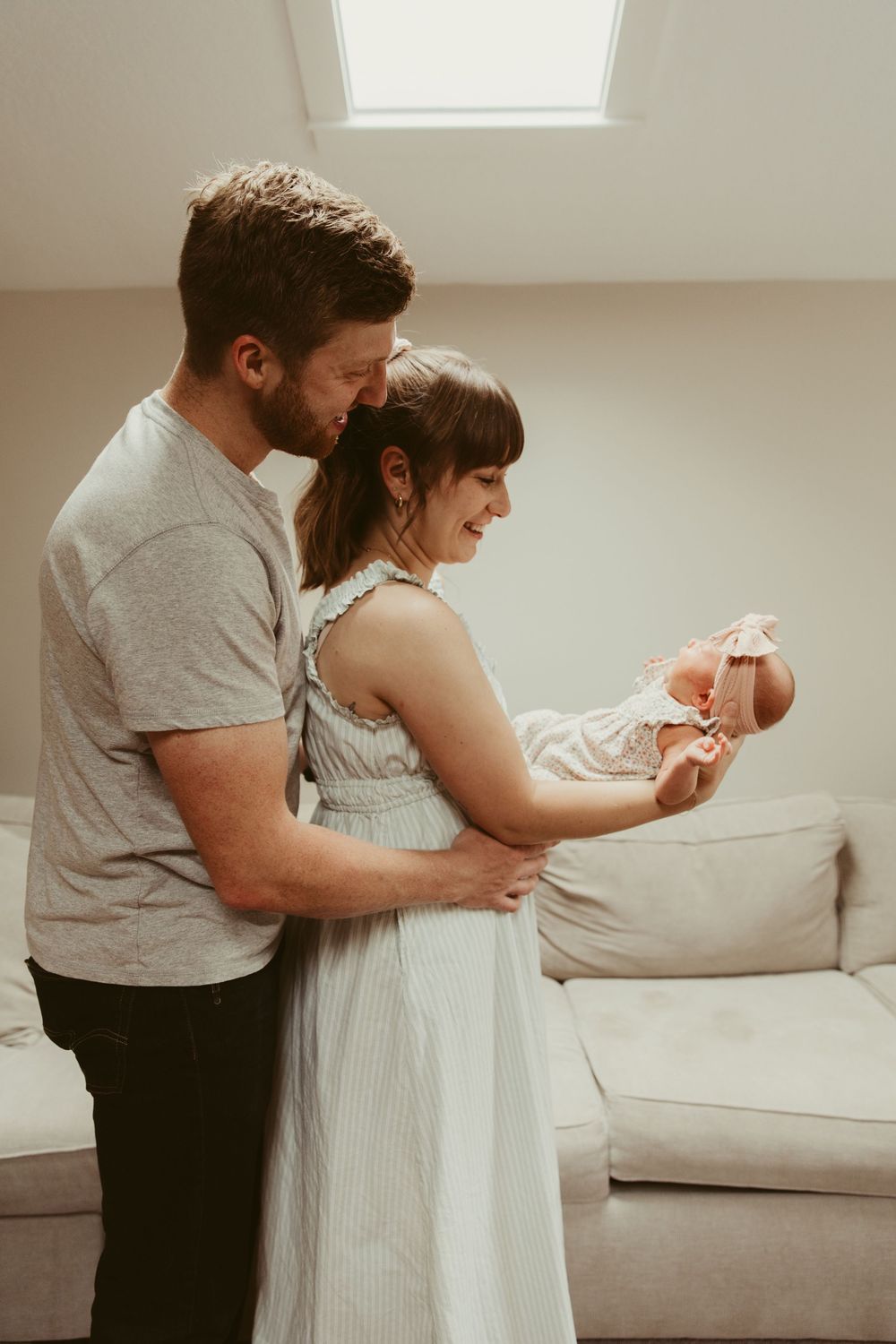 A couple in a bright room holds their newborn baby while standing near a white couch under a skylight.