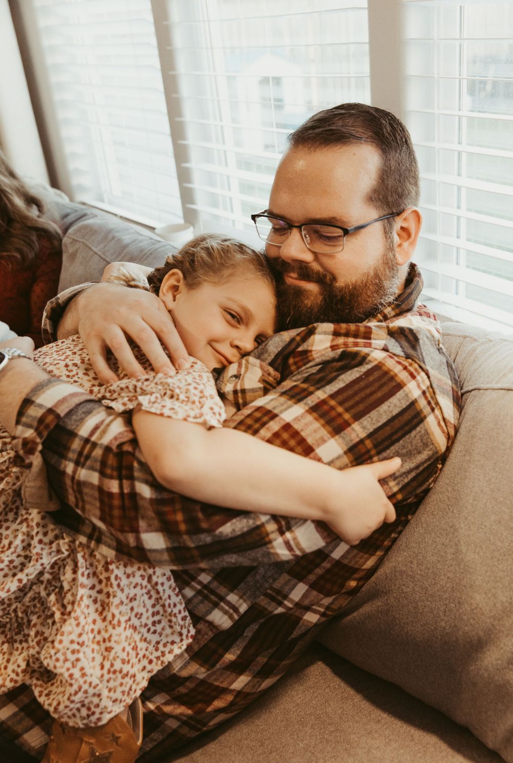 A warm embrace shared between two people on a gray couch, with natural light streaming through nearby windows.