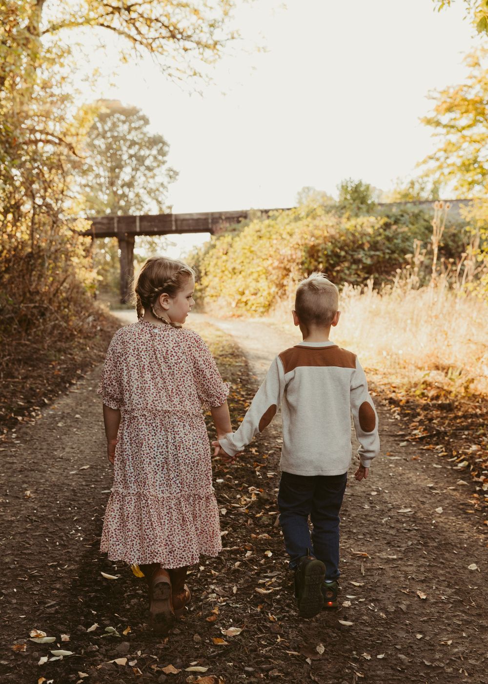 Two young children walk hand in hand down a sunlit autumn path with golden leaves falling around them.