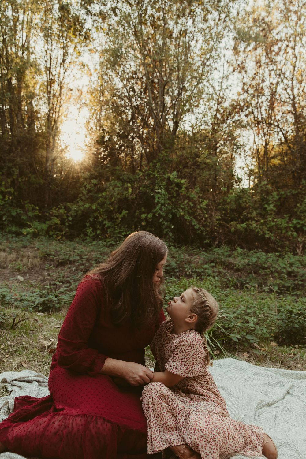 Mother and child share tender moments on a white blanket outdoors during golden hour sunset.