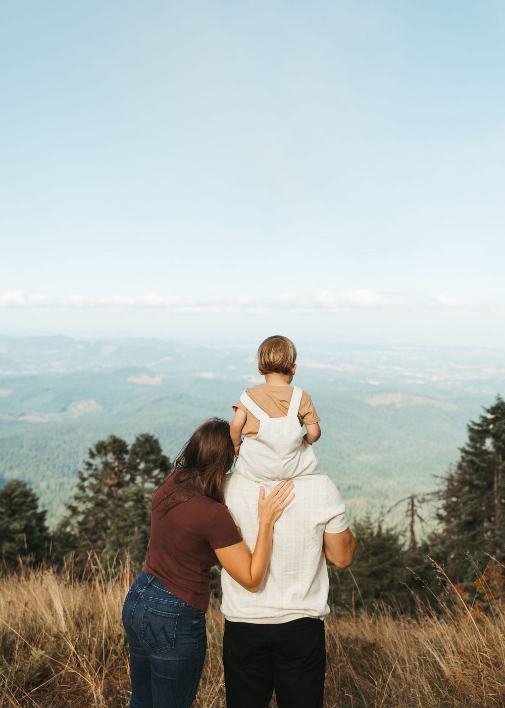 Family of three enjoying a scenic mountain overlook, with child sitting on father's shoulders and mother beside them.