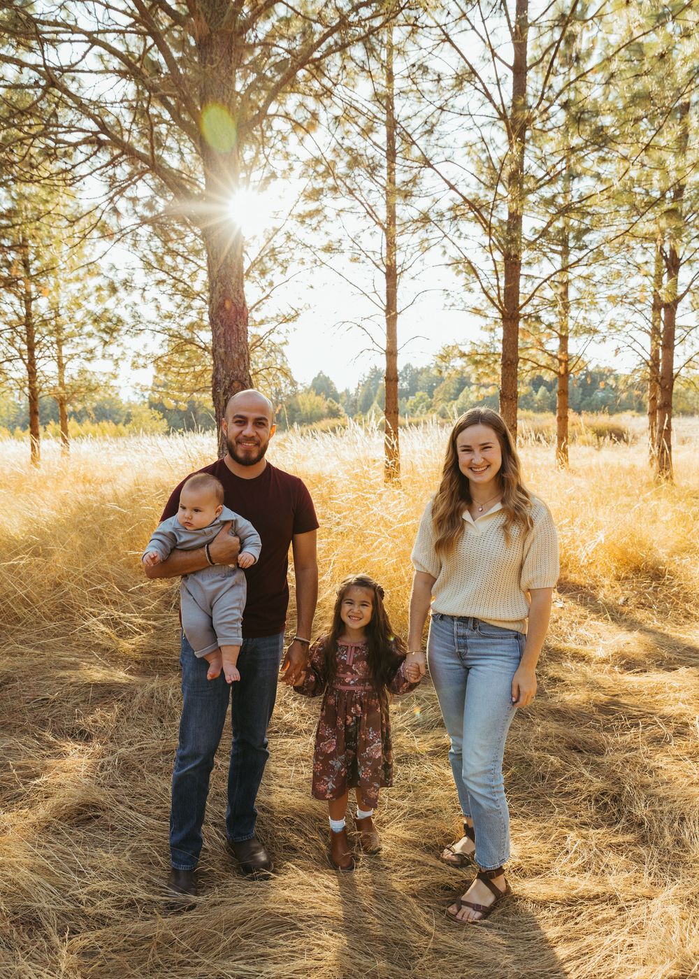 A family stands together in a golden field with tall pine trees and sunlight streaming through the branches.
