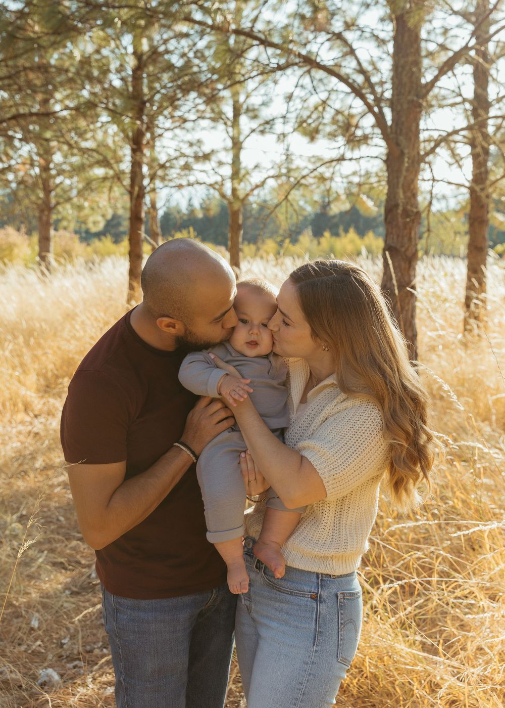 Family shares tender moments together in a golden field with tall pine trees during sunset.