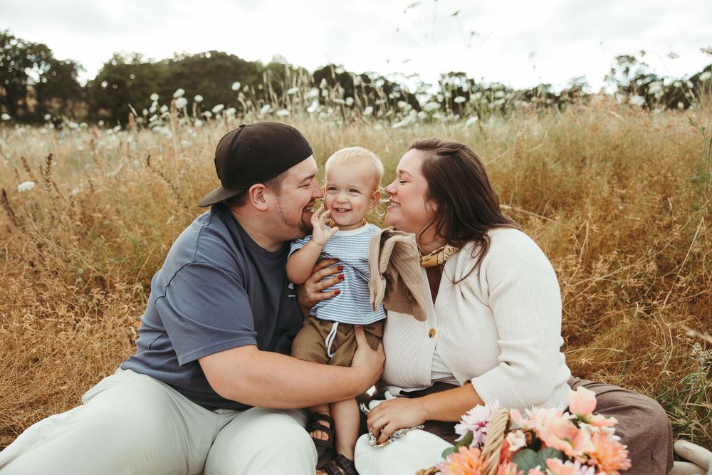 A happy young family with a baby sits in a golden field, sharing a tender moment with flowers nearby.