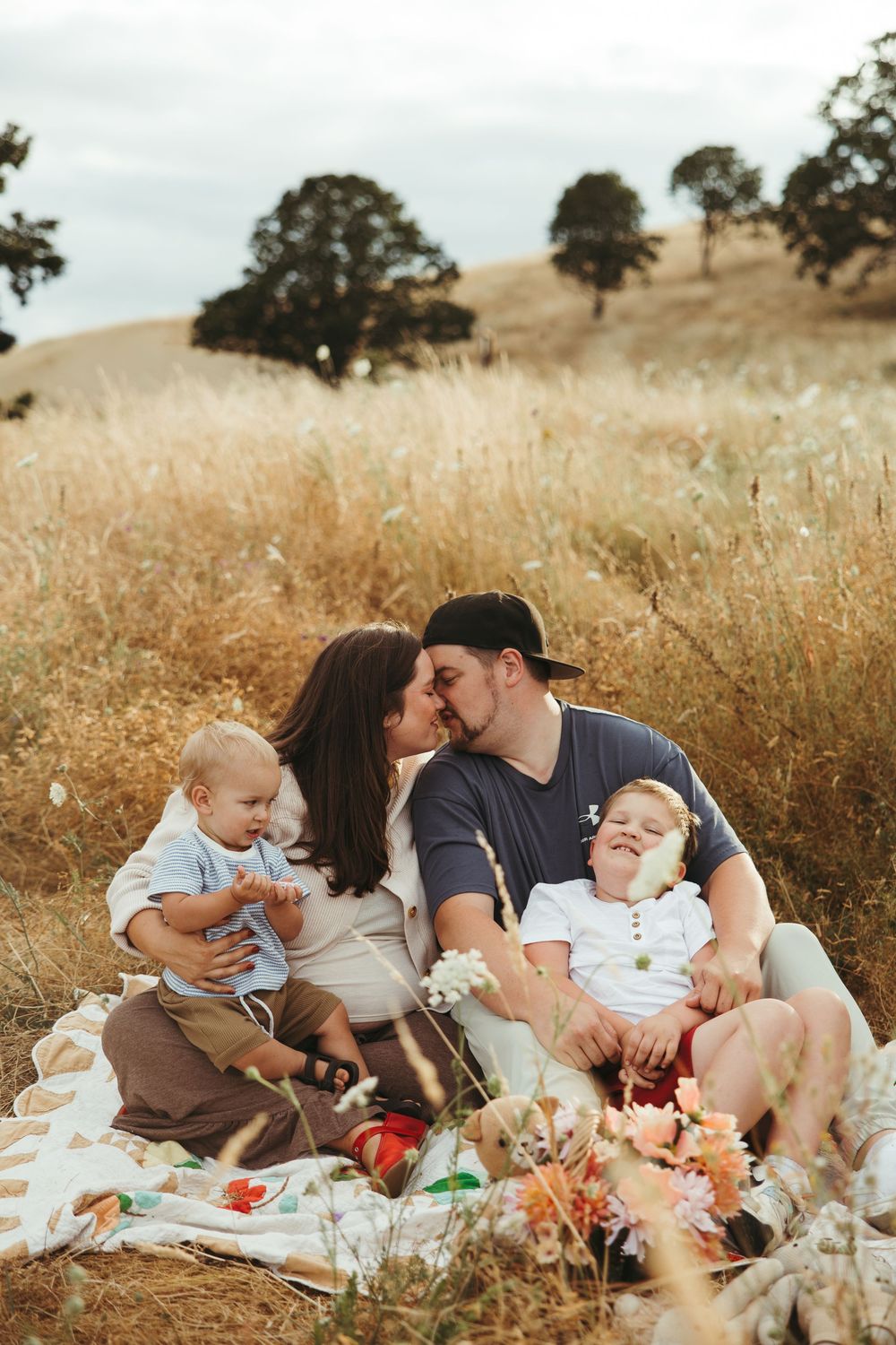 A young family sits on a blanket in a golden field, parents sharing a kiss while holding their two toddlers.