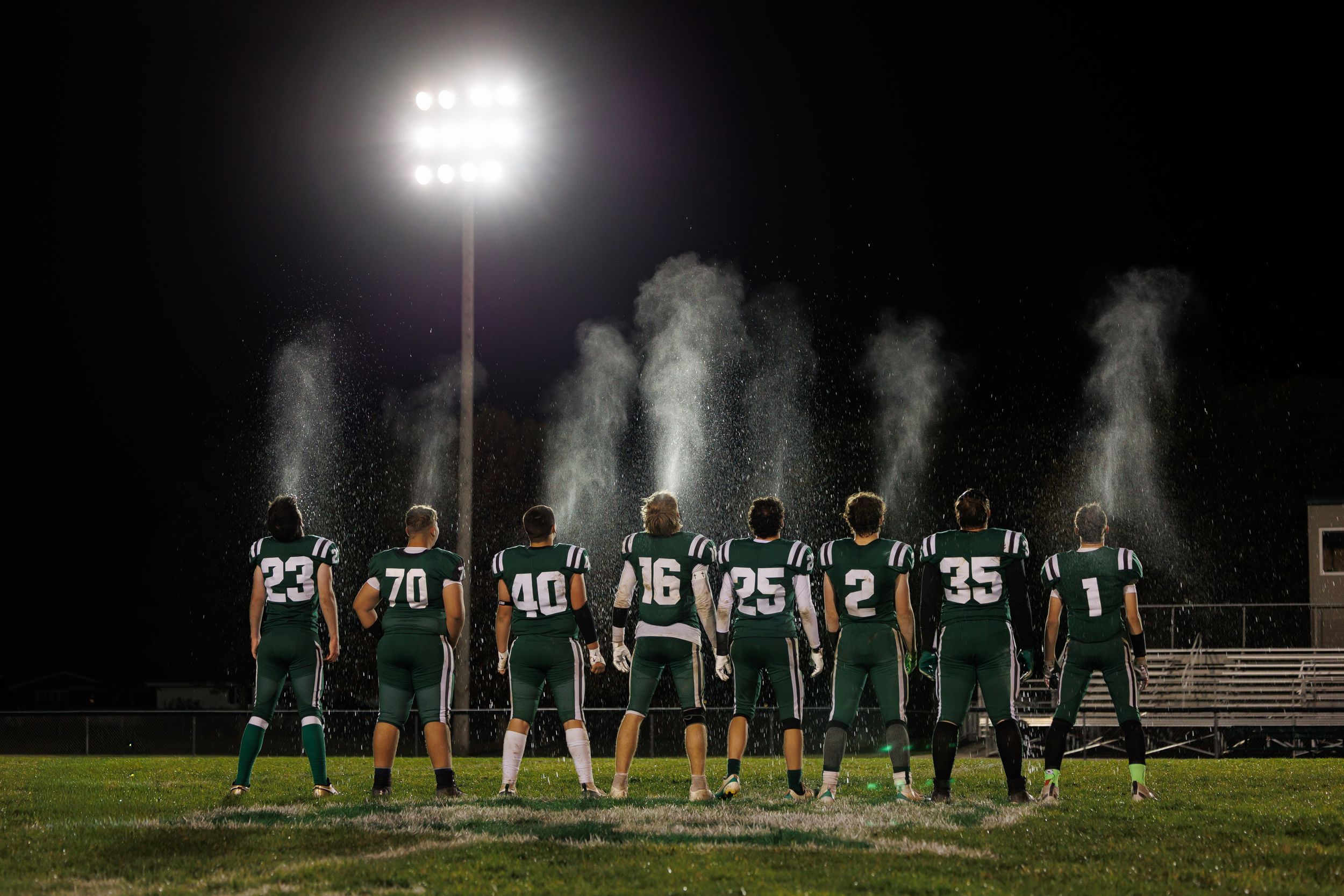 Football players stand silhouetted against bright stadium lights during a nighttime game.