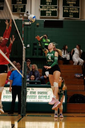 Volleyball players compete at the net during an intense match in a high school gymnasium with championship banners visible.