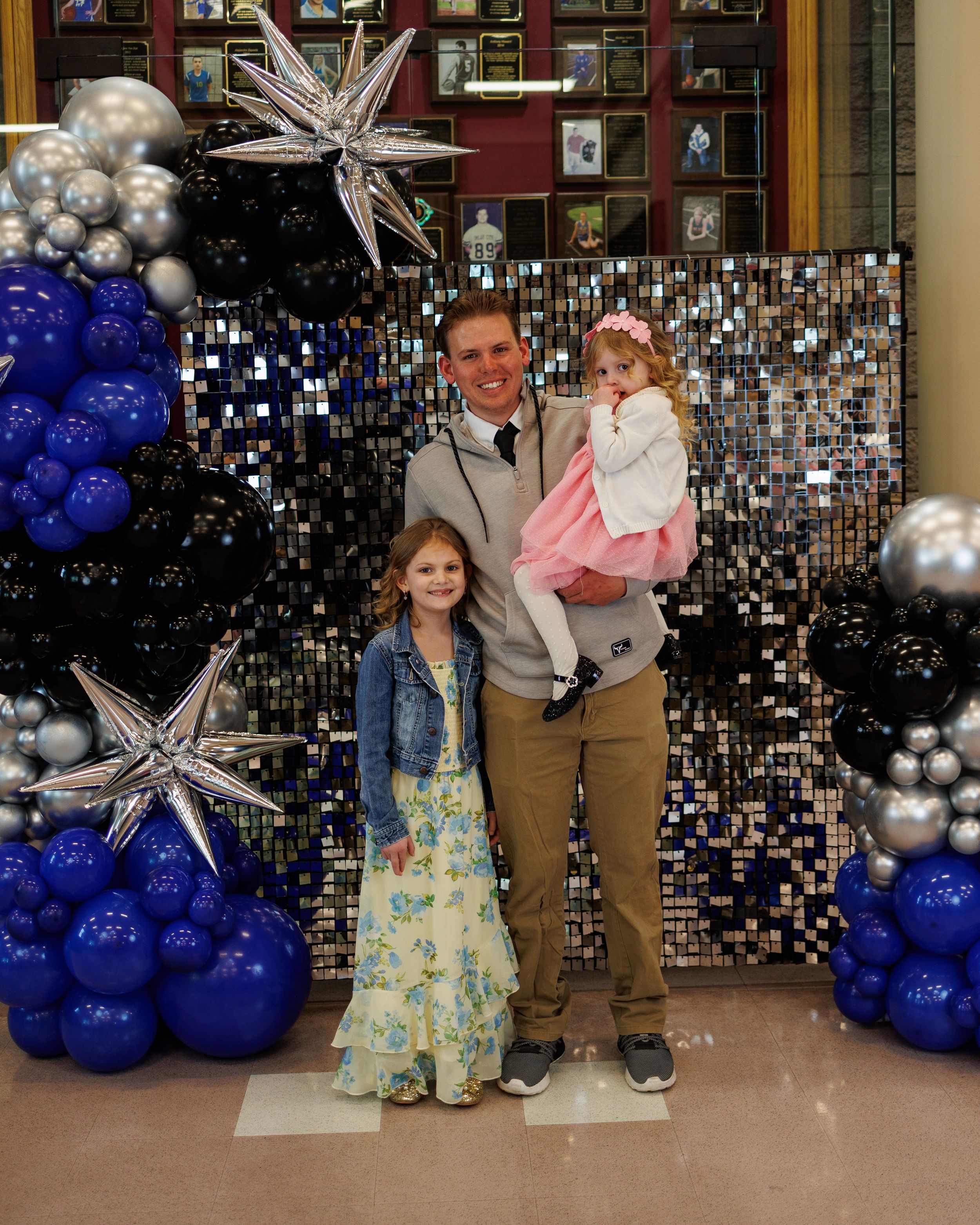 Man posing with two young girls in front of blue, black, and silver balloon decorations with star balloons.