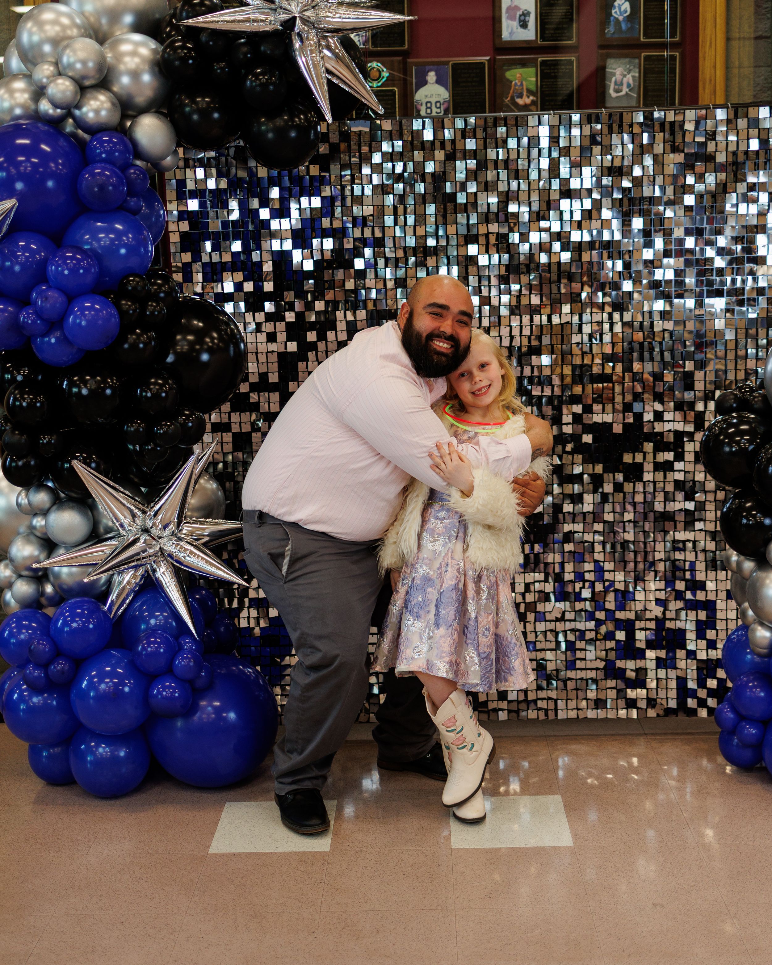 A man and young girl pose together in front of a shimmery backdrop with blue, black, and silver star balloons.