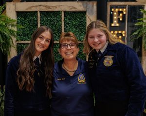 Students in FFA blue jackets pose together at an event with decorative greenery and lights in the background.