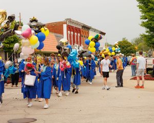 A panoramic view of graduates in blue robes gathering outside with colorful balloons during a graduation ceremony.